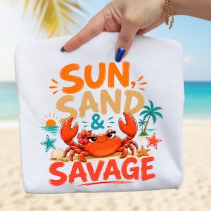 May include: White tote bag with a colorful beach-themed graphic. The design features the words "SUN, SAND & SAVAGE" in orange and red, along with a cartoon crab, starfish, and palm trees. The background suggests a beach setting.