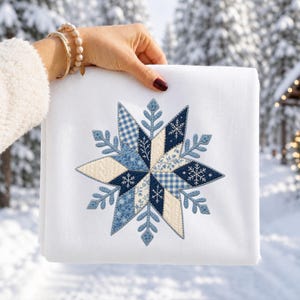 May include: A white fabric item featuring a large, central embroidered snowflake design. The snowflake is composed of various blue and cream-colored patches with patterns including gingham, floral, and cable knit textures, accented by small white snowflakes.
