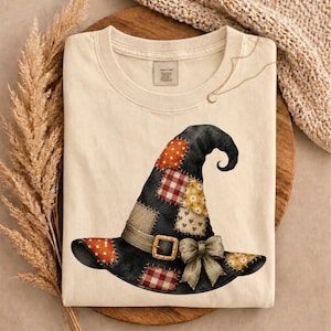 May include: A cream-colored t-shirt featuring a graphic of a black witch hat decorated with various patchwork patterns including orange polka dots, red gingham, yellow floral, and heart prints, finished with a buckle and a bow.