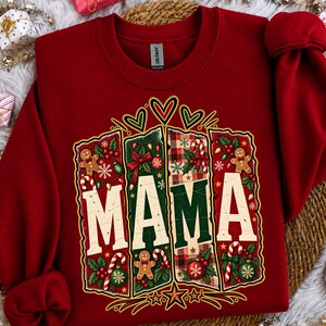 Könnte beinhalten: Ein dunkelrotes Sweatshirt mit Rundhalsausschnitt und einem zentralen Aufdruck des Wortes MAMA in großen weißen Buchstaben. Das Design besteht aus vier vertikalen Feldern, die mit Lebkuchenmännern, Zuckerstangen, roten Kugeln und Stechpalmen verziert sind.