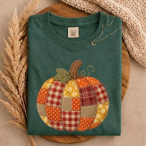 May include: A dark green crew neck t-shirt featuring a large central graphic of a pumpkin made from a patchwork of various fabrics including red and white gingham, orange polka dots, yellow floral patterns, and solid tan textures.