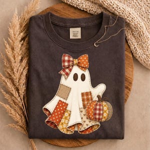 May include: A dark gray t-shirt featuring a central graphic of a ghost made of various fabric patches in orange, brown, and cream colors. The ghost wears a plaid and polka dot bow and holds a small pumpkin with a matching patchwork pattern.