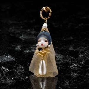 Puede incluir: Un pendiente único con una cara de muñeca con un gorro azul y un lazo dorado. Una gran perla cuelga debajo de la cara y un anillo dorado remata el pendiente. El pendiente está sobre un fondo oscuro.