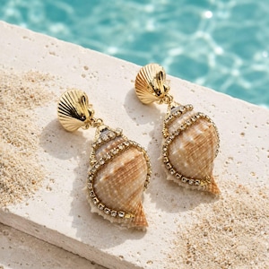 Peut inclure: Une paire de boucles d'oreilles pendantes composées de coquillages naturels en spirale de couleur beige, ornées de capuchons en métal doré et de petits strass transparents incrustés le long des nervures. Fermoir à tige en métal doré.