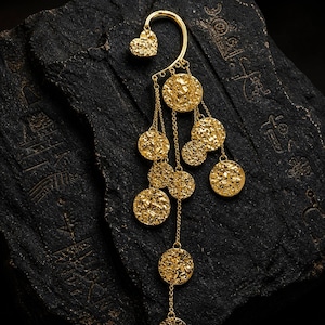 May include: A gold-colored ear cuff with a heart-shaped stud and dangling textured coin charms. The ear cuff curves around the ear and features multiple chains with circular pendants. The jewelry is set against a dark, textured background.