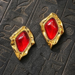 May include: A pair of gold-toned earrings with a red gemstone center. The earrings have an irregular, organic shape and are set against a dark, textured background with engraved symbols. The gemstones have a faceted appearance.