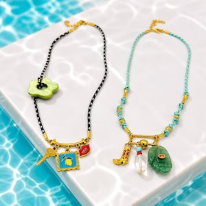 May include: Two necklaces with gold-toned accents and colorful charms. One features a black and white cord with a green flower and charms including a key, lemon, and lips. The other has a turquoise cord with a boot, a clear vial, and a green leaf.