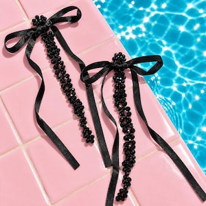 May include: Two black beaded hair accessories with satin ribbon bows. The beaded strands are long and dangle from the bows. The accessories are on a pink tiled surface next to a pool of blue water.