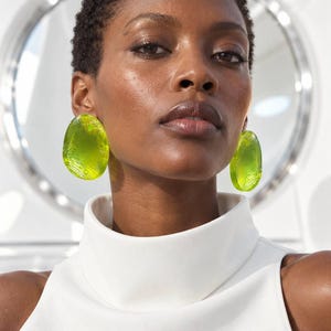 May include: A pair of large, oval-shaped earrings in a vibrant, translucent lime green color. The earrings feature a textured, rippled surface that catches the light, creating a glossy appearance. They are worn against a plain white high-neck top.