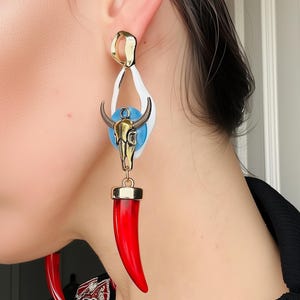 Puede incluir: Pendientes dorados con un acento blanco y azul, con un colgante de calavera de toro y un cuerno rojo grande. Los pendientes son una pieza audaz, que muestra un diseño único.