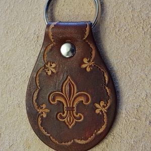 May include: A brown leather keychain with a fleur-de-lis design. The keychain has a silver metal ring.