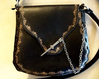 Peut inclure: Un sac à bandoulière en cuir noir avec une chaîne argentée et un fermoir métallique décoratif. Le sac est orné de broderies dorées sur les bords.