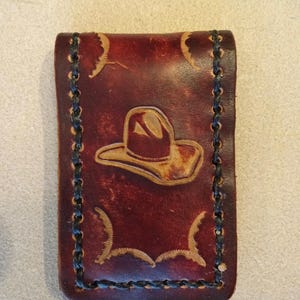 May include: A brown leather wallet with a black stitched border and a tooled design of a cowboy hat.