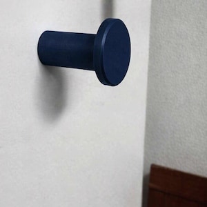May include: A dark navy blue wall hook with a cylindrical base and a flat, circular front face. The hook is mounted horizontally, extending outward from a light-colored wall surface, featuring a smooth, matte finish throughout its design.