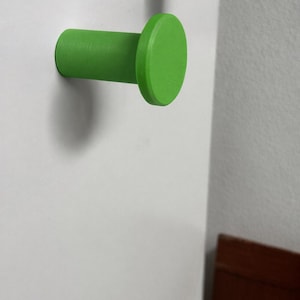 May include: A bright lime green wall hook with a cylindrical base and a flat, circular front face. The hook is mounted horizontally, extending outward from a flat, white vertical surface, featuring a smooth, matte finish throughout its design.