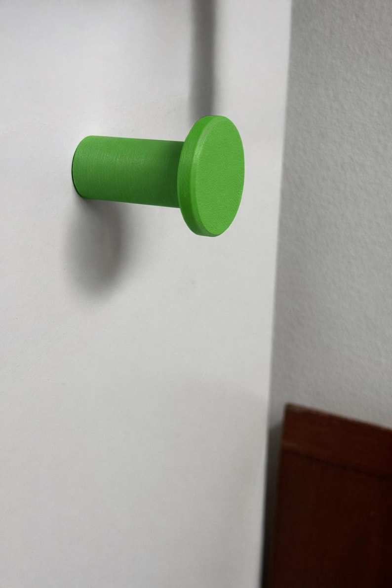 May include: A bright lime green wall hook with a cylindrical base and a flat, circular front face. The hook is mounted horizontally, extending outward from a flat, white vertical surface, featuring a smooth, matte finish throughout its design.