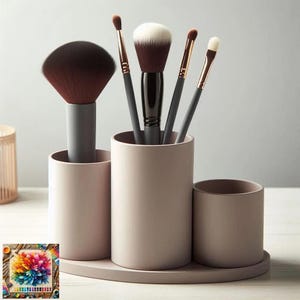 May include: A collection of makeup brushes in a pale pink holder. The brushes feature dark brown and white bristles with grey and gold handles. The holder has three cylindrical sections and a small, round section, all on a matching tray. A colourful painting is in the corner.