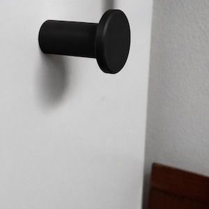 May include: A matte black wall hook featuring a cylindrical body and a flat, circular front face. The hook is mounted horizontally, extending outward from the surface, providing a simple, functional design for hanging various items.