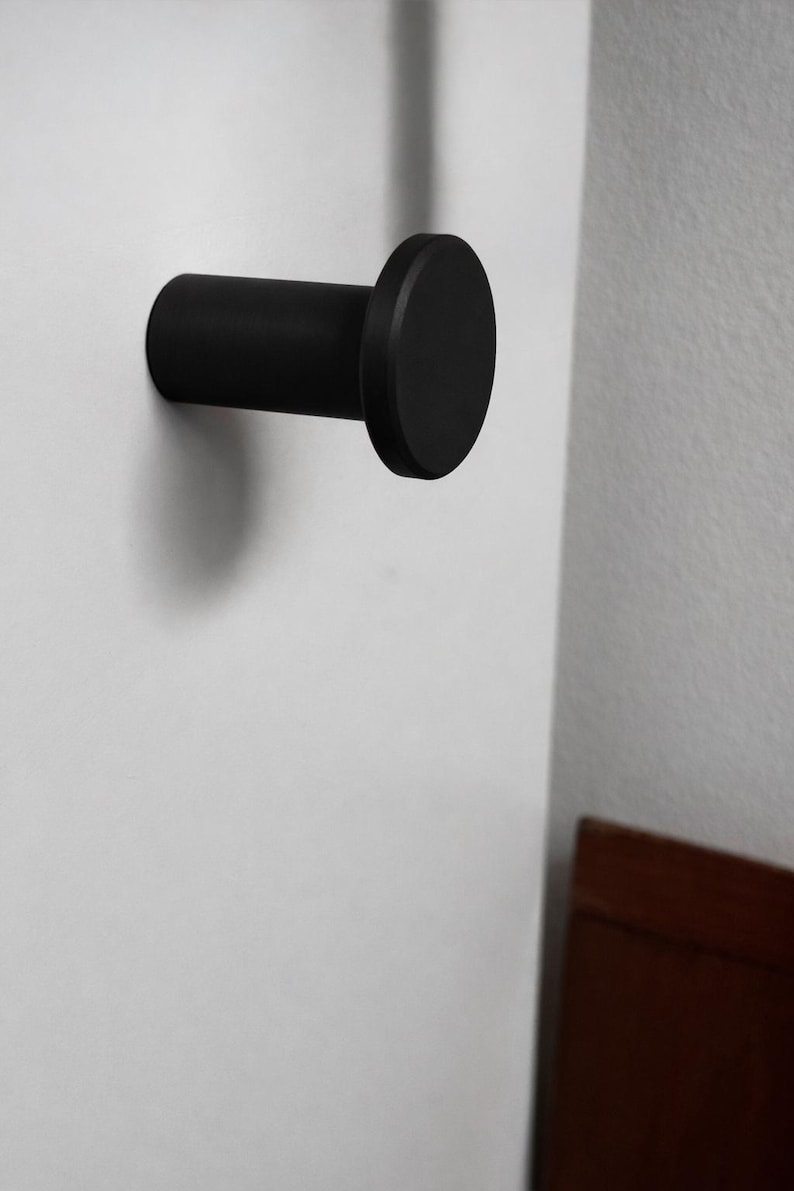 May include: A matte black wall hook featuring a cylindrical body and a flat, circular front face. The hook is mounted horizontally, extending outward from the surface, providing a simple, functional design for hanging various items.