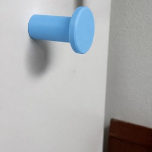 May include: A light blue wall hook with a cylindrical base and a flat circular front face. The hook is mounted horizontally, extending outward from a flat surface, featuring a smooth, matte finish and a simple, functional geometric design.