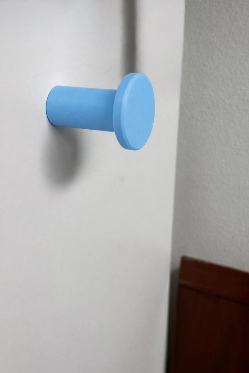 May include: A light blue wall hook with a cylindrical base and a flat circular front face. The hook is mounted horizontally, extending outward from a flat surface, featuring a smooth, matte finish and a simple, functional geometric design.