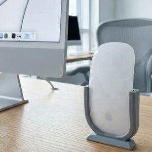 May include: A white Apple Magic Mouse in a grey holder on a wooden desk. The mouse, with the Apple logo, sits beside a silver computer monitor. A grey office chair is in the background.