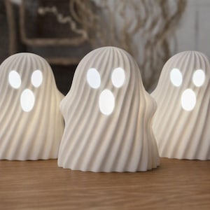 May include: Three white ghost-shaped lamps with a ribbed, swirling texture. Each lamp features two oval eyes and an oval mouth that emit a bright, soft white light, creating a glowing effect against the matte surface of the ghost figures.