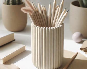 Könnte beinhalten: Ein zylindrischer Stiftehalter mit vertikaler Rippenstruktur in hellem Beige. Der Halter ist mit mehreren gespitzten Holzstiften und zwei flauschigen, hellbraunen Zierzweigen gefüllt. Das Objekt hat eine glatte, matte Oberfläche.