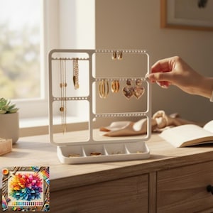 May include: A white jewellery organiser with multiple compartments and earring holders. The organiser is on a wooden surface, displaying various gold-toned earrings and necklaces. A hand is reaching towards the organiser, adding an earring. A small tray at the base holds rings.