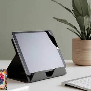 May include: A dark grey tablet stand supporting a tablet with a blank screen. A white keyboard and a potted plant are also visible. A colourful painting is in the lower left corner.