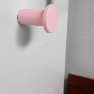 May include: A cylindrical wall hook with a flat, circular front face, finished in a solid matte light pink color. The hook extends horizontally from the mounting surface, featuring a smooth, uniform texture and a simple, functional design.