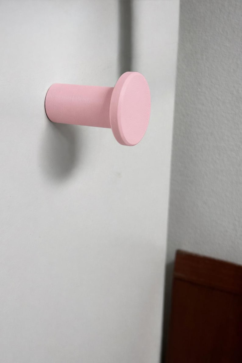 May include: A cylindrical wall hook with a flat, circular front face, finished in a solid matte light pink color. The hook extends horizontally from the mounting surface, featuring a smooth, uniform texture and a simple, functional design.