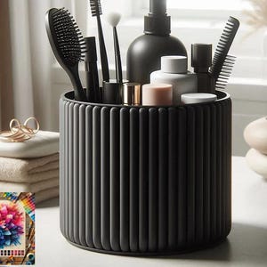 May include: A dark grey, ribbed cylindrical container holding various grooming tools, including brushes and combs. The container sits on a white surface, with folded towels and artwork in the background.