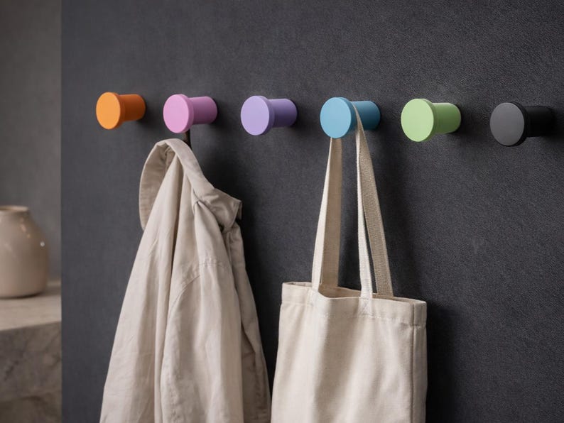 May include: A row of six cylindrical wall hooks in orange, pink, lavender, light blue, light green, and black. A beige jacket hangs from the pink hook, and a plain off-white canvas tote bag hangs from the light blue hook on the dark wall.