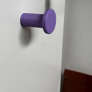 May include: A purple wall-mounted hook with a cylindrical base and a flat, circular front face. The hook is attached to a smooth white surface, extending outward with a simple, uniform design that features a matte finish and clean edges.