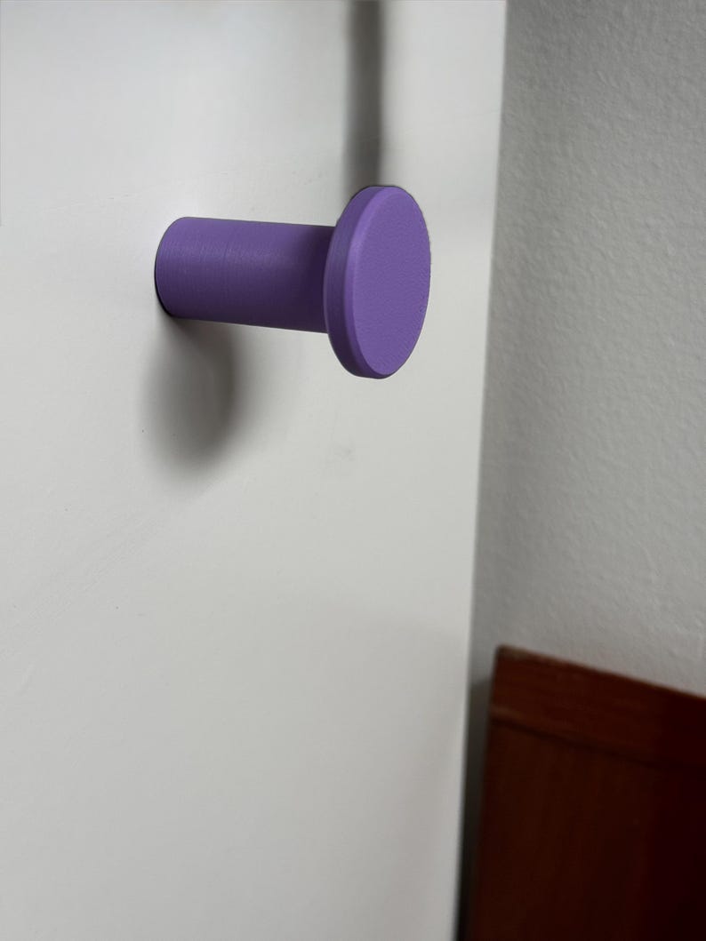 May include: A purple wall-mounted hook with a cylindrical base and a flat, circular front face. The hook is attached to a smooth white surface, extending outward with a simple, uniform design that features a matte finish and clean edges.