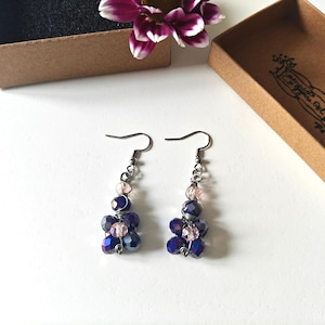Puede incluir: Un par de pendientes colgantes con cuentas de cristal azul oscuro y rosa dispuestas en un diseño floral. Los pendientes cuelgan de ganchos plateados y se muestran cerca de cajas marrones abiertas y una flor morada.