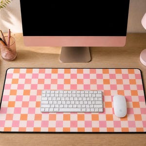 以下が含まれることがあります： ピンクとオレンジのチェック柄のマウスパッド、白いキーボード、白いマウスを備えたデスクセットアップ。ピンクのコンピューターモニター、ピンクのデスクランプ、ピンクのノートも表示されます。デスクは木製です。