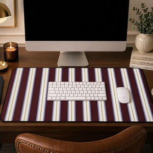 May include: A rectangular desk mat with a striped pattern in burgundy, white, and light blue. A white keyboard and mouse sit on the mat, which is placed on a dark wooden desk. A computer monitor is in the background.