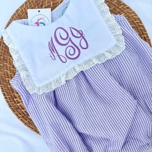 May include: A lavender and white striped seersucker romper with a white collar. The collar is trimmed with eyelet lace and features a purple monogram. The romper is designed for children.