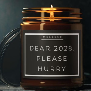 Peut inclure: Une bougie en verre brun avec une étiquette noire sur laquelle est inscrit "Cher 2028, S'il te plaît, dépêche-toi". La bougie est allumée et la flamme est visible.