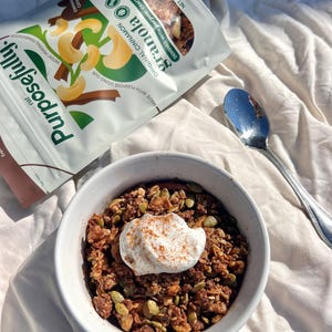 May include: A bowl of granola topped with cream and cinnamon, next to a spoon. A bag of Purposefully Original Cinnamon Granola is also visible. The bag shows cashews and the word "granola". The bowl is on a white surface.