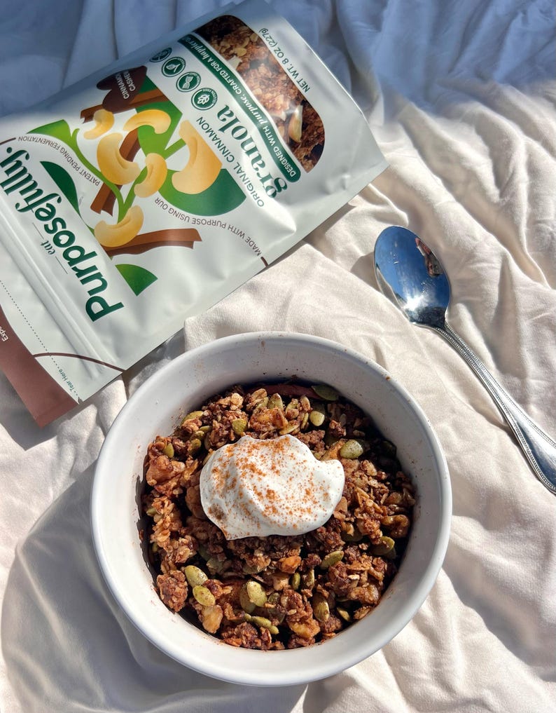 May include: A bowl of granola topped with cream and cinnamon, next to a spoon. A bag of Purposefully Original Cinnamon Granola is also visible. The bag shows cashews and the word "granola". The bowl is on a white surface.