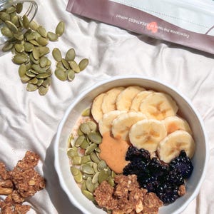 May include: A white bowl containing a breakfast of sliced bananas, blueberries, pumpkin seeds, granola, and peanut butter. A bag of Purposefull granola is in the background. The text on the bag reads "Experience the taste of GREATNESS within!"