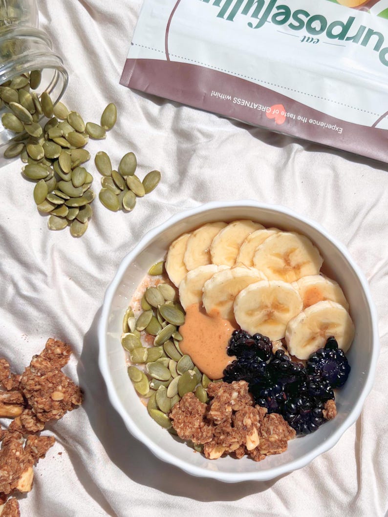 May include: A white bowl containing a breakfast of sliced bananas, blueberries, pumpkin seeds, granola, and peanut butter. A bag of Purposefull granola is in the background. The text on the bag reads "Experience the taste of GREATNESS within!"