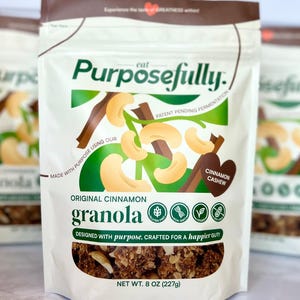 May include: A white packet of "Purposefully" Original Cinnamon Granola. The packaging displays cashews and cinnamon sticks. The text reads "Designed with purpose, crafted for a happier gut!" and "Net Wt. 227g".