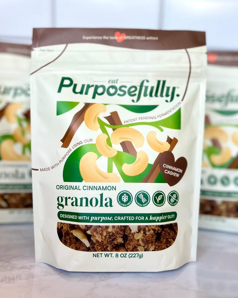 May include: A white packet of "Purposefully" Original Cinnamon Granola. The packaging displays cashews and cinnamon sticks. The text reads "Designed with purpose, crafted for a happier gut!" and "Net Wt. 227g".