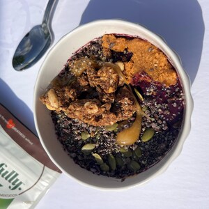 May include: A white bowl filled with a colourful breakfast. The bowl contains a dark base, topped with granola, seeds, and a swirl of peanut butter. A spoon rests beside the bowl, and a bag of food is partially visible.