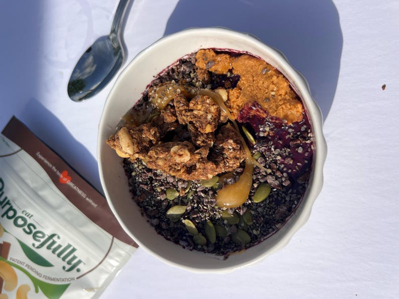 May include: A white bowl filled with a colourful breakfast. The bowl contains a dark base, topped with granola, seeds, and a swirl of peanut butter. A spoon rests beside the bowl, and a bag of food is partially visible.