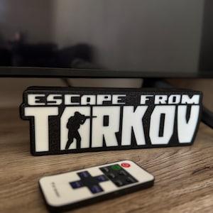 Pode incluir: Placa preta e branca com a inscrição "ESCAPE FROM TARKOV" e a silhueta de um soldado. Um controle remoto branco com botões pretos está na frente. A placa está em uma superfície de madeira.